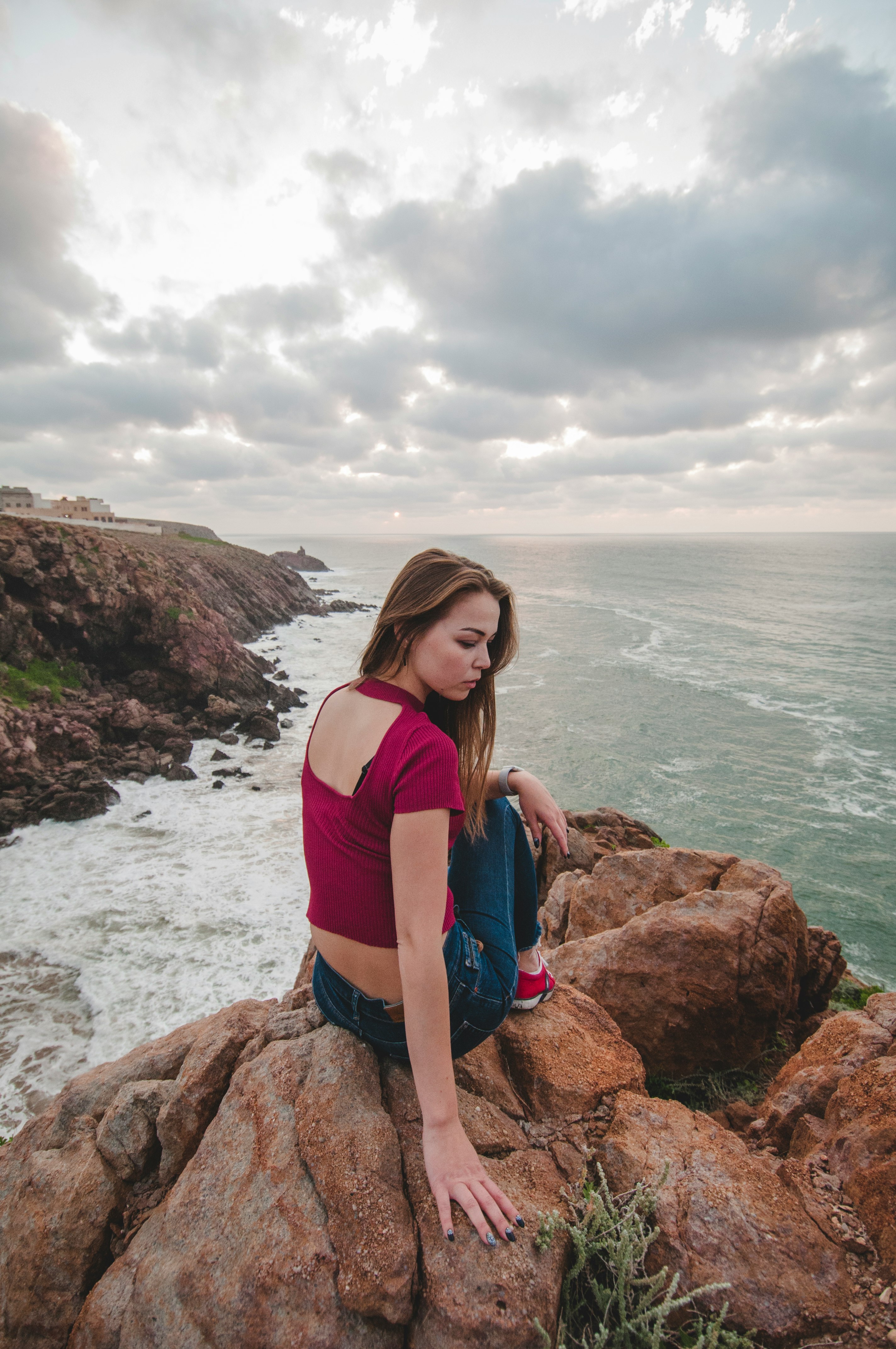 woman in red tank top and blue denim jeans sitting on brown rock formation near sea